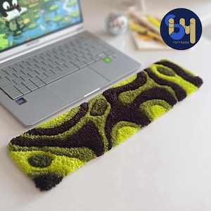 Könnte beinhalten: Ein rechteckiger, getufteter Teppich mit einem einzigartigen Muster aus Dunkelbraun und verschiedenen Grüntönen. Der Teppich liegt auf einer weißen Oberfläche in der Nähe eines Laptops. Das Logo von TUFTRUGS ist sichtbar.