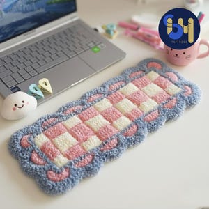 Puede incluir: Una alfombra rectangular, afelpada, con un patrón de tablero de ajedrez de cuadrados rosas y blancos, enmarcada por un borde azul con acentos rosas en forma de corazón. La alfombra está colocada sobre una superficie blanca cerca de un portátil y una taza rosa.