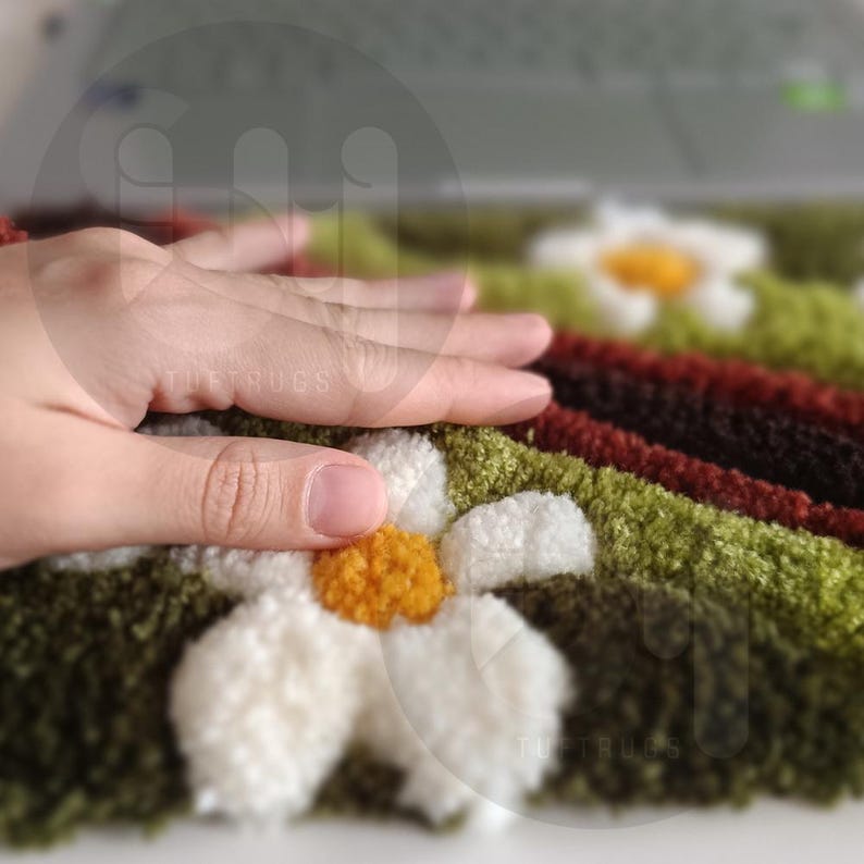 May include: Close-up of a tufted rug with a daisy design. The rug features white daisy petals with yellow centers, set against a green and brown background. The text "TUFT RUGS" is visible. The rug appears soft and textured.