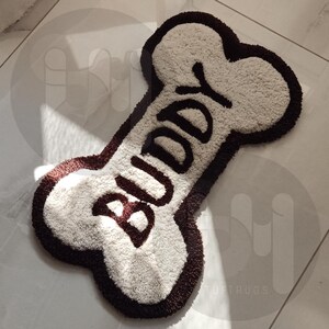 May include: A white and brown dog bone shaped rug with the word "Buddy" embroidered in brown lettering.