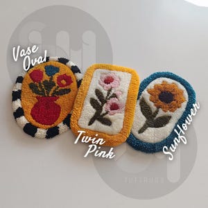May include: Three tufted rugs with floral designs. The first rug is oval shaped and features a vase with flowers in red, yellow, and green. The second rug is rectangular and features pink flowers on a white background. The third rug is oval shaped and features a yellow sunflower on a blue background. The rugs are labeled "Vase Oval", "Twin Pink", and "Sunflower".