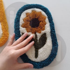 May include: A white oval rug with a blue border and a yellow sunflower with a brown center and green leaves in the center.