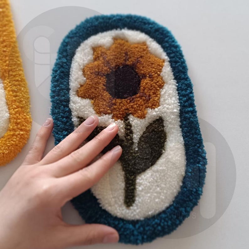 May include: A white oval rug with a blue border and a yellow sunflower with a brown center and green leaves in the center.