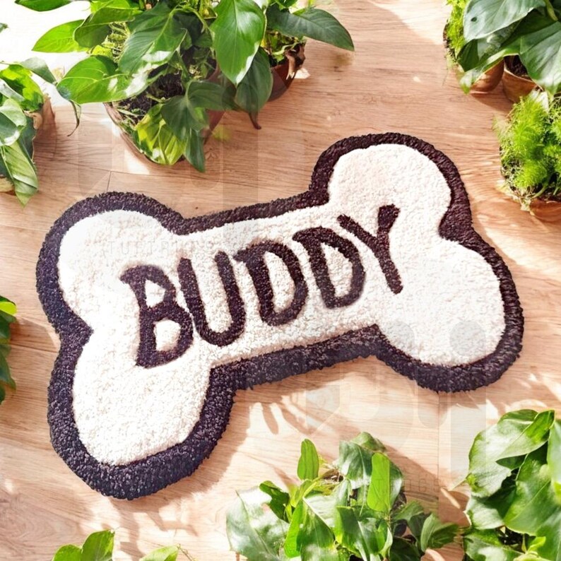 May include: A white and brown dog bone shaped rug with the word "BUDDY" written in brown on the top. The rug is on a wooden floor.