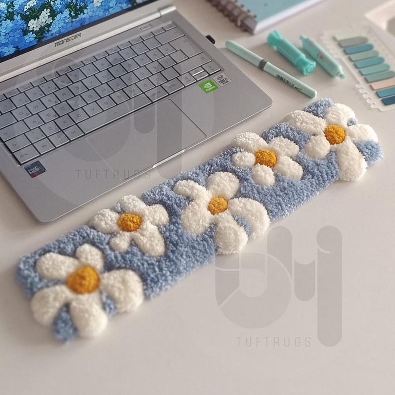 May include: A light blue, rectangular rug with a daisy pattern. The rug features white daisy petals with yellow centers, set against a textured blue background. The rug is placed on a white surface near a laptop.