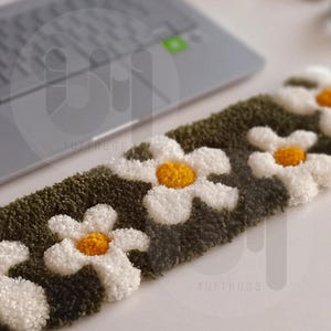 May include: A close-up of a green and white tufted rug with a daisy pattern. The rug has a fluffy texture and is made with a variety of colors. The text "tuftrugs" is visible in the lower right corner of the image.
