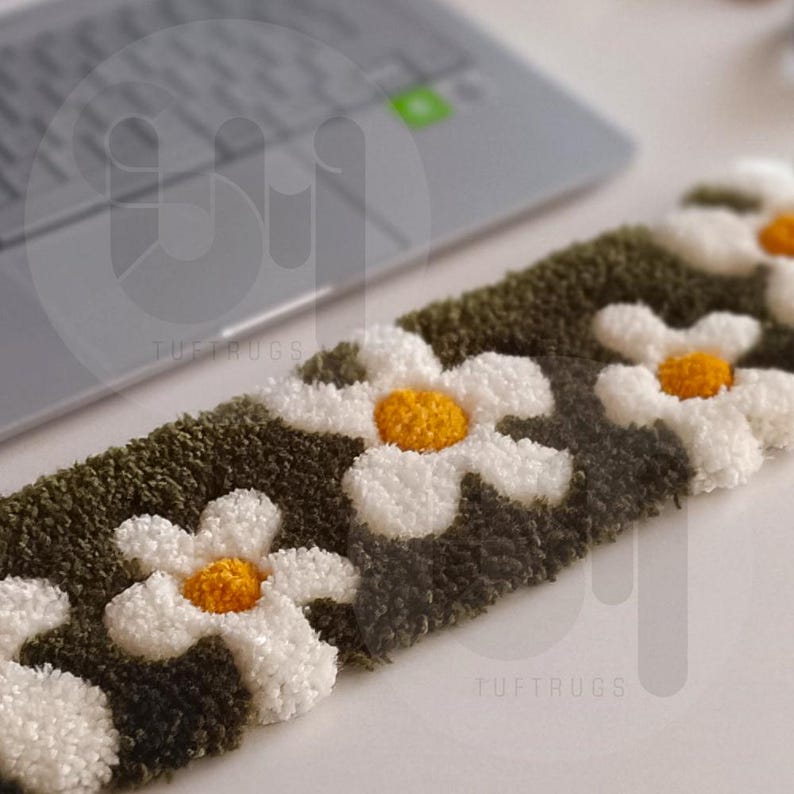 May include: A close-up of a green and white tufted rug with a daisy pattern. The rug has a fluffy texture and is made with a variety of colors. The text "tuftrugs" is visible in the lower right corner of the image.