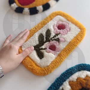 May include: A yellow framed rug with a white background and a floral design featuring two pink flowers and green leaves.