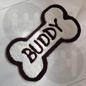 May include: White dog bone shaped rug with a brown border and the word "Buddy" written in brown lettering.