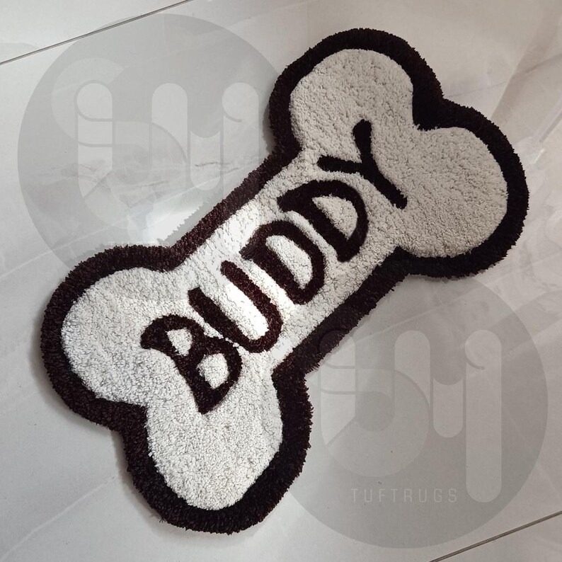 May include: White dog bone shaped rug with a brown border and the word "Buddy" written in brown lettering.