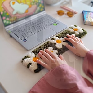 May include: A green and white tufted rug with a daisy pattern. The rug is placed on a white desk with a laptop and a pair of sunglasses.