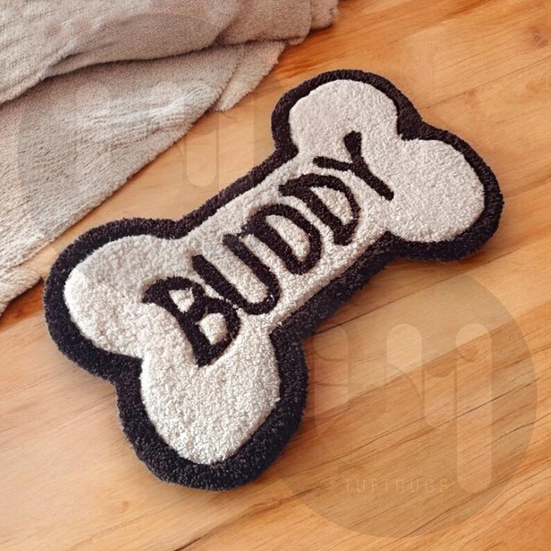 May include: A white bone-shaped doormat with a black border and the word "Buddy" written in black cursive lettering.