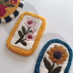 May include: Three embroidered patches with floral designs. The first patch is yellow with a black and white stripe border and features a red flower. The second patch is yellow with a white background and features two pink flowers. The third patch is teal with a white background and features a yellow sunflower.