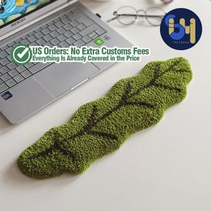May include: A green leaf-shaped rug with dark brown veins, placed on a white surface. The rug is made of textured material. The text "TUFTRUGS" is visible in the image.