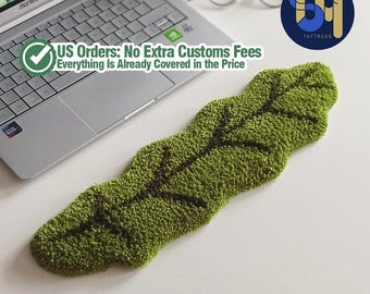 May include: A green leaf-shaped rug with dark brown veins, placed on a white surface. The rug is made of textured material. The text "TUFTRUGS" is visible in the image.