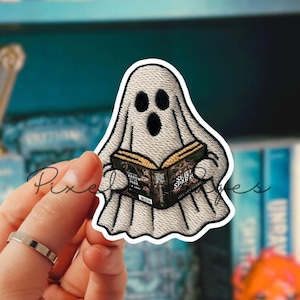 May include: A white ghost sticker with black eyes and mouth, holding a book with the title "SHOE". The sticker is held in front of a bookshelf filled with books. The ghost is reading a book.