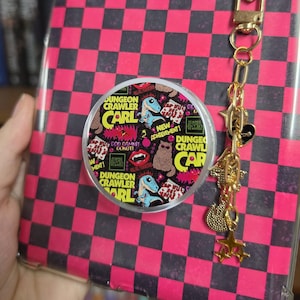 Puede incluir: Una funda rectangular para tableta con patrón de tablero de ajedrez rosa y negro, que presenta un agarre circular desplegable decorado con gráficos de Dungeon Crawler Carl y un llavero de metal dorado con dijes de corazón y estrella.