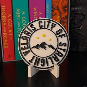 May include: A white and black circular coaster with the text "VELARIS CITY OF STARLIGHT" and a mountain and star design. The coaster is displayed on a white stand, with colorful books in the background.