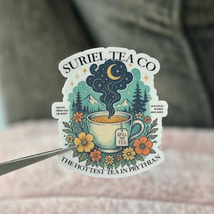 May include: A sticker featuring a whimsical design for Suriel Tea Co. The illustration includes a teacup with steam rising to a night sky with a crescent moon, surrounded by flowers and trees. Text includes "Spill the Tea" and "The Hottest Tea in Prythian."