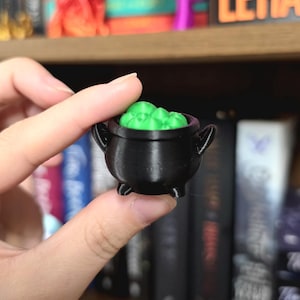 May include: A miniature black cauldron with three small feet and two side handles, filled with a bright green bubbling liquid texture. The object is constructed from a dark material with visible horizontal print lines throughout the surface.