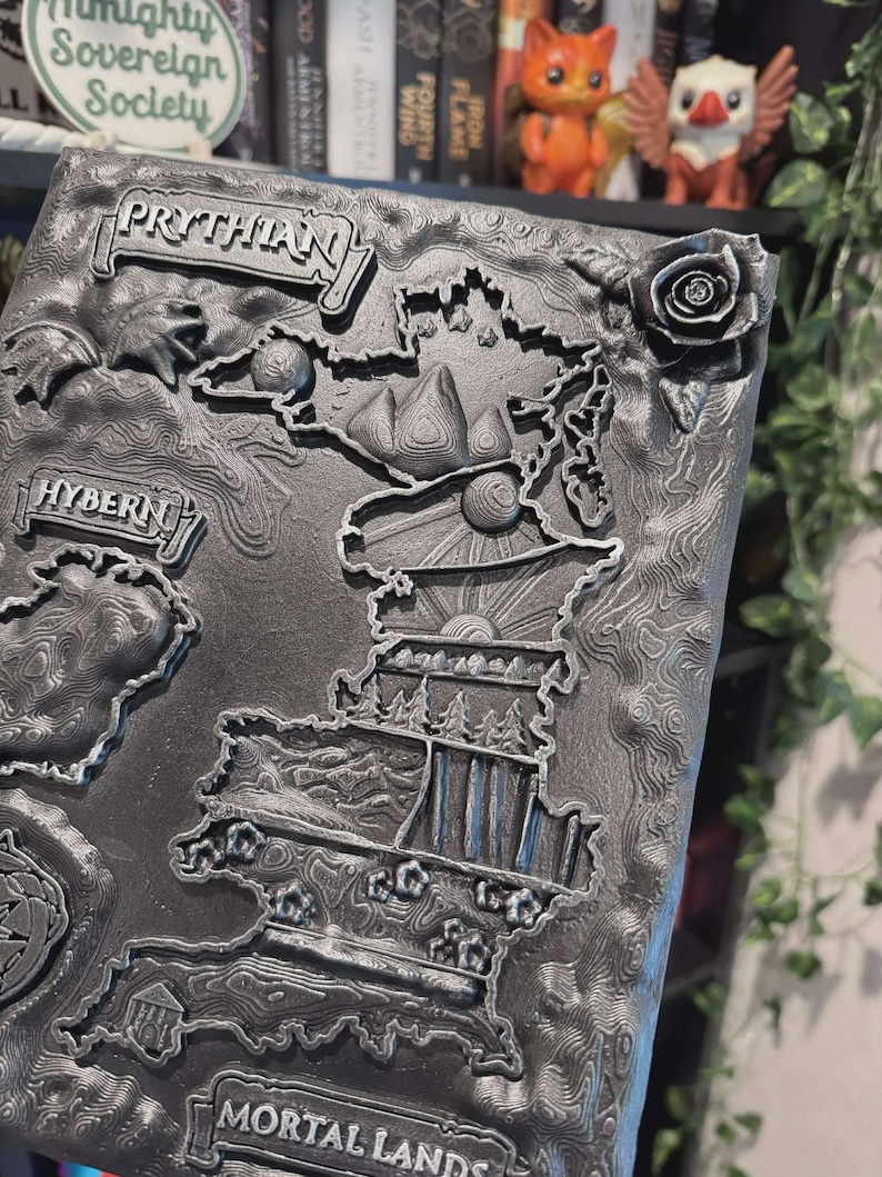 May include: A detailed, gray, 3D-printed map featuring the words "PRYTHIAN", "HYBERN", and "MORTAL LANDS". The map includes raised relief details of landmasses, mountains, and a rose. The background has a textured pattern.