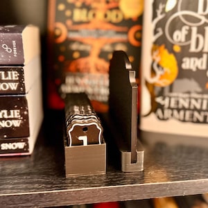 May include: A black, decorative book stand with a metal base, holding a stack of small, brown tags with white lettering. The tags are in a small, brown box. Books with titles like "Kylie Snow" and "Blood and Ash" are in the background.