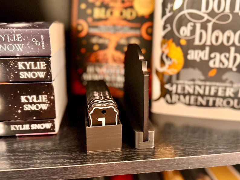 May include: A black, decorative book stand with a metal base, holding a stack of small, brown tags with white lettering. The tags are in a small, brown box. Books with titles like "Kylie Snow" and "Blood and Ash" are in the background.