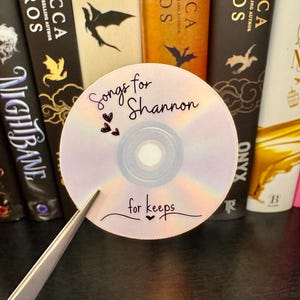 May include: A CD with the text "Songs for Shannon" and "for keeps" in black script. The CD is held by tweezers and is in front of a row of books with various titles and colors.
