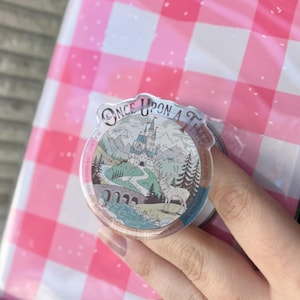 Pode incluir: Um suporte de telefone redondo e transparente com uma paisagem de conto de fadas com um castelo, montanhas e um veado. O texto "ONCE UPON A TIME" está arqueado na parte superior. Sobre um fundo de xadrez rosa e branco.