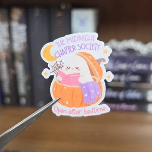K&ouml;nnte beinhalten: Ein gestanzter Aufkleber mit einem wei&szlig;en Geist, der in einem orangefarbenen K&uuml;rbis sitzt und ein rosa Buch liest. Das Design enth&auml;lt einen gelben Halbmond, ein Spinnennetz und eine violette Decke. Der Text lautet: THE MIDNIGHT CHAPTER SOCIETY und Open after bedtime.
