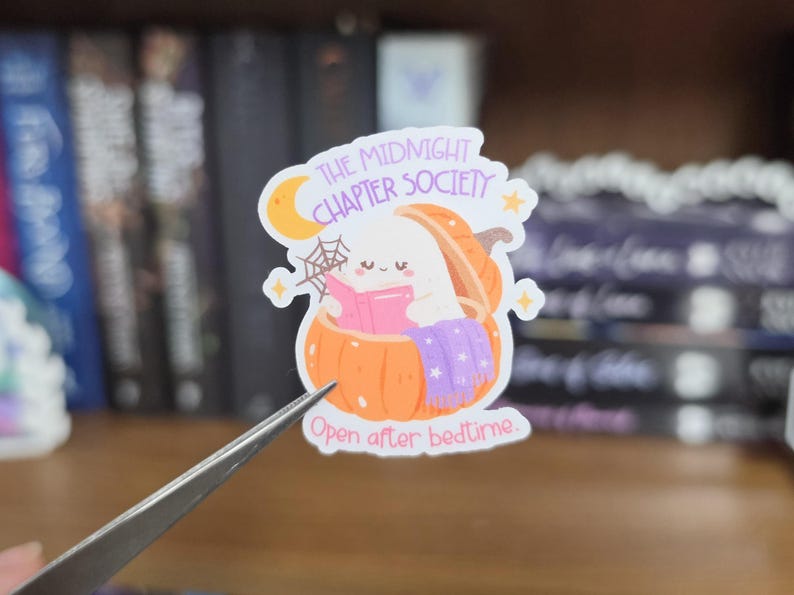 K&ouml;nnte beinhalten: Ein gestanzter Aufkleber mit einem wei&szlig;en Geist, der in einem orangefarbenen K&uuml;rbis sitzt und ein rosa Buch liest. Das Design enth&auml;lt einen gelben Halbmond, ein Spinnennetz und eine violette Decke. Der Text lautet: THE MIDNIGHT CHAPTER SOCIETY und Open after bedtime.