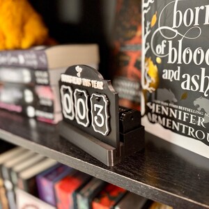 May include: A black and white book tracker with the text "Read This Year" and the numbers "003". The tracker is on a shelf with books, including one titled "Born of Blood and Ash".
