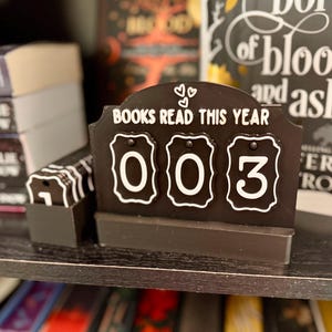 May include: A black and white book counter displays "BOOKS READ THIS YEAR" with the number "003". The counter has a decorative top with two hearts. The counter is on a shelf with books in the background.