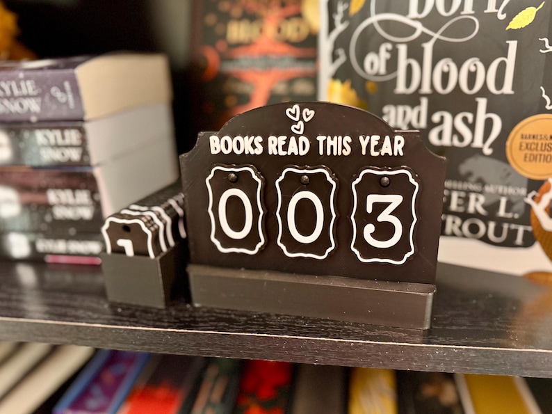 May include: A black and white book counter displays "BOOKS READ THIS YEAR" with the number "003". The counter has a decorative top with two hearts. The counter is on a shelf with books in the background.