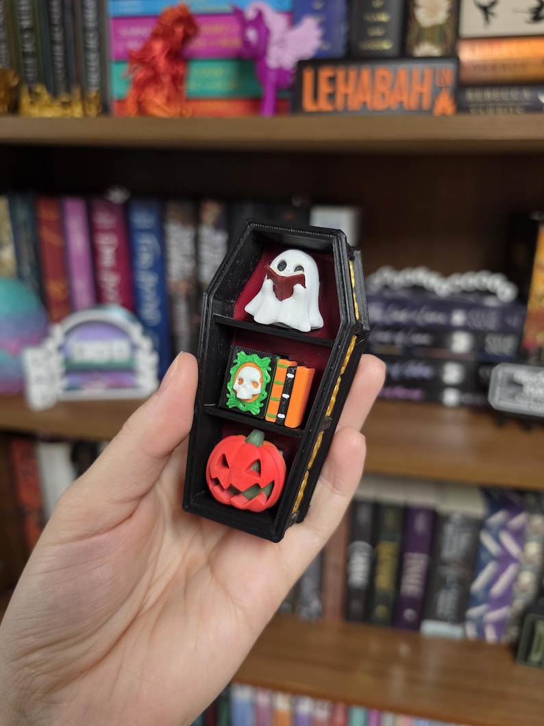 May include: A black coffin-shaped shelf with three tiers. The top shelf holds a white ghost reading a book. The middle shelf contains a green framed skull and two orange books. The bottom shelf features a red jack-o'-lantern with a green stem.