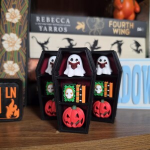 May include: A black coffin-shaped shelf with a dark red interior featuring three levels. The shelves hold miniature white ghosts reading books, green books with skull covers, orange books, and small orange jack-o'-lantern pumpkins.