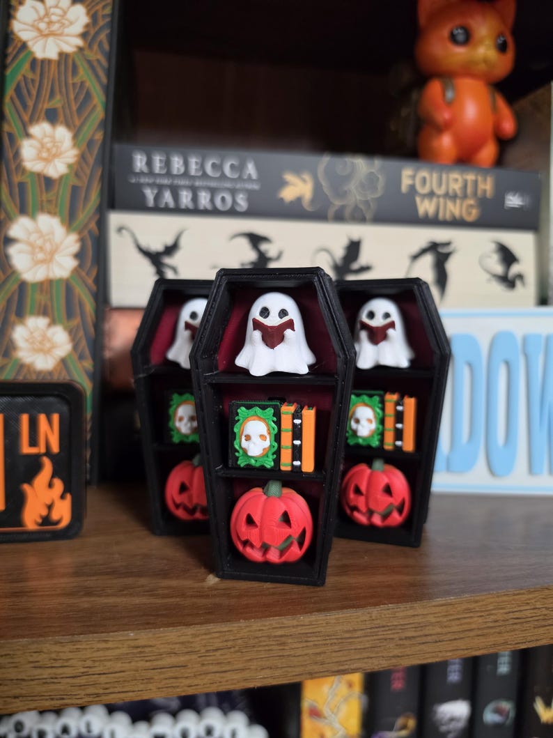 May include: A black coffin-shaped shelf with a dark red interior featuring three levels. The shelves hold miniature white ghosts reading books, green books with skull covers, orange books, and small orange jack-o'-lantern pumpkins.