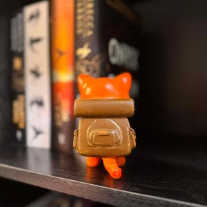 May include: A 3D-printed orange cat figurine with a brown backpack. The cat has a small tail and is standing on a shelf in front of a row of books. The figurine is a unique decorative item.
