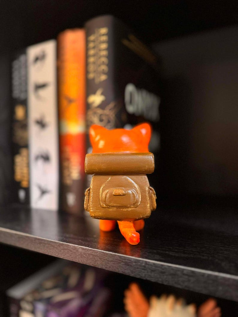 May include: A 3D-printed orange cat figurine with a brown backpack. The cat has a small tail and is standing on a shelf in front of a row of books. The figurine is a unique decorative item.