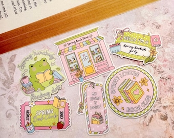 May include: A collection of colorful stickers with a spring book theme. Designs include a frog reading, a bookstore, a ticket, a bookmark, and a circular sticker with the text "Spring Lovers Book Club". The stickers have pastel colors and floral accents.