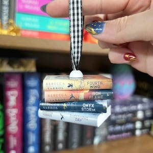May include: A miniature book ornament featuring a stack of four titles by Rebecca Yarros: Fourth Wing, Iron Flame, Onyx Storm, and Threshing Day. The stack is suspended by a black and white gingham ribbon attached to a small white bead at the top.