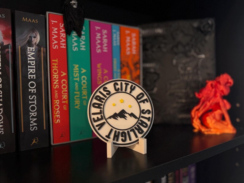 May include: A white and black round sign with the text "VELARIS CITY OF STARLIGHT" and a mountain graphic, displayed on a white stand. The sign is on a shelf with books and an orange sculpture.