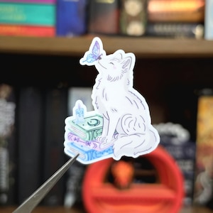May include: A die-cut sticker featuring a white fox sitting on a stack of three books. A small blue candle sits on the top book, and a purple butterfly rests on the fox's nose. The illustration uses soft blue, purple, and white color tones.