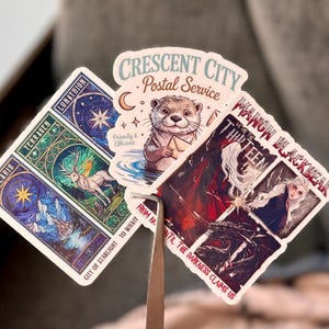 May include: A collection of fantasy-themed stickers. One features an otter in a postal uniform, another has a deer and celestial imagery, and a third depicts a woman with long white hair. Text includes "Crescent City Postal Service" and "Manon Blackbeak".
