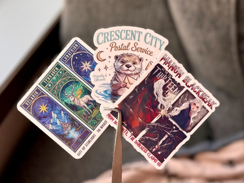 May include: A collection of fantasy-themed stickers. One features an otter in a postal uniform, another has a deer and celestial imagery, and a third depicts a woman with long white hair. Text includes "Crescent City Postal Service" and "Manon Blackbeak".