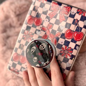 Könnte beinhalten: Eine Handyhülle mit einem schwarz-weißen Schachbrettmuster, roten Kirschen, Schleifen und Herzen. Ein Pop-Socket mit schwarzem Hintergrund und weißen Totenköpfen ist an der Handyhülle befestigt.
