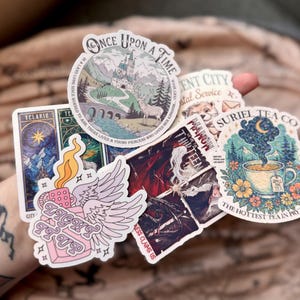 May include: A collection of colorful stickers featuring various designs. One sticker reads "Once Upon a Time" with a castle illustration. Other stickers include text like "Telaris", "Light It Up", and "Suriel Tea Co."
