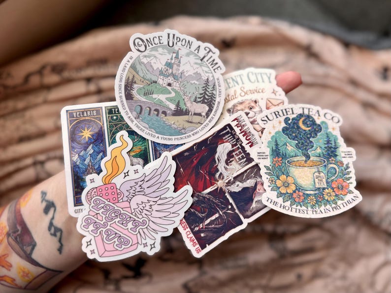 May include: A collection of colorful stickers featuring various designs. One sticker reads "Once Upon a Time" with a castle illustration. Other stickers include text like "Telaris", "Light It Up", and "Suriel Tea Co."