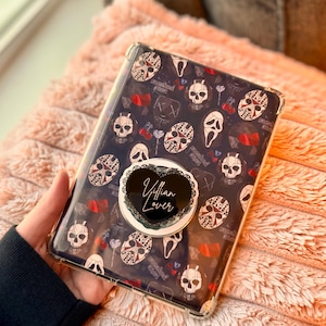 以下が含まれることがあります： 白頭蓋骨、赤いアクセント、テキストのパターンが施された、濃い青色の背景の透明な携帯電話ケース。 「Villain Lover」の文字が入った黒いハート型の電話グリップがケースに取り付けられています。