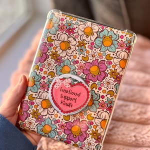 May include: A clear tablet case with a colorful floral pattern featuring orange, purple, and blue flowers. A pink heart-shaped pop socket with the text "Emotional Support Kindle" is attached to the case. The case is held in a person's hand.